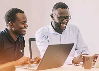 two Kenyan men smiling and and talking as they work on laptops and drink out of a mug